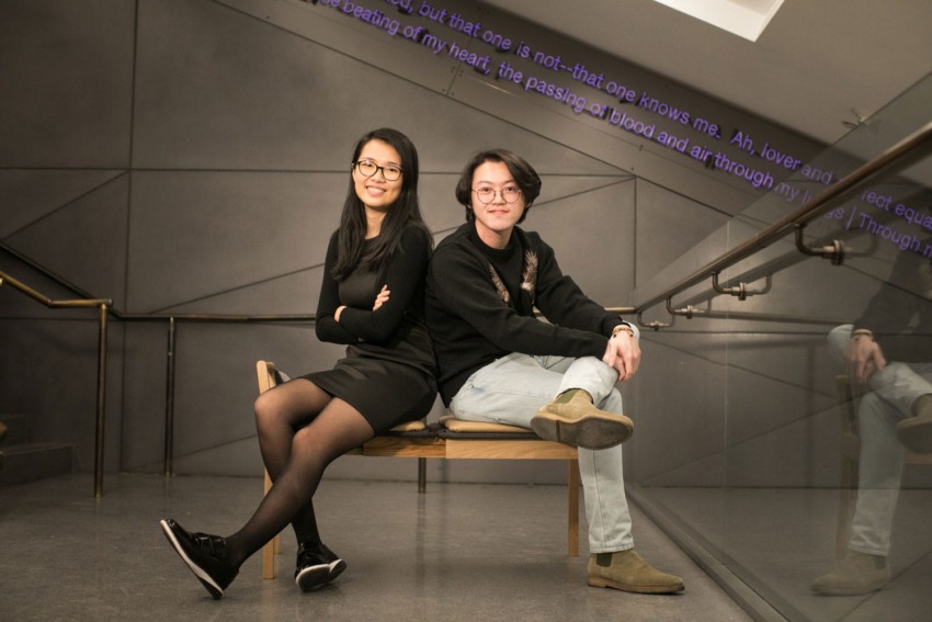 Two people sitting back-to-back on a bench in a modern, angular room with a metal railing and text projected on the ceiling.