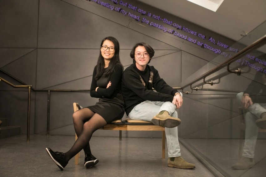 Two people sitting back-to-back on a bench in a modern, angular room with a metal railing and text projected on the ceiling.