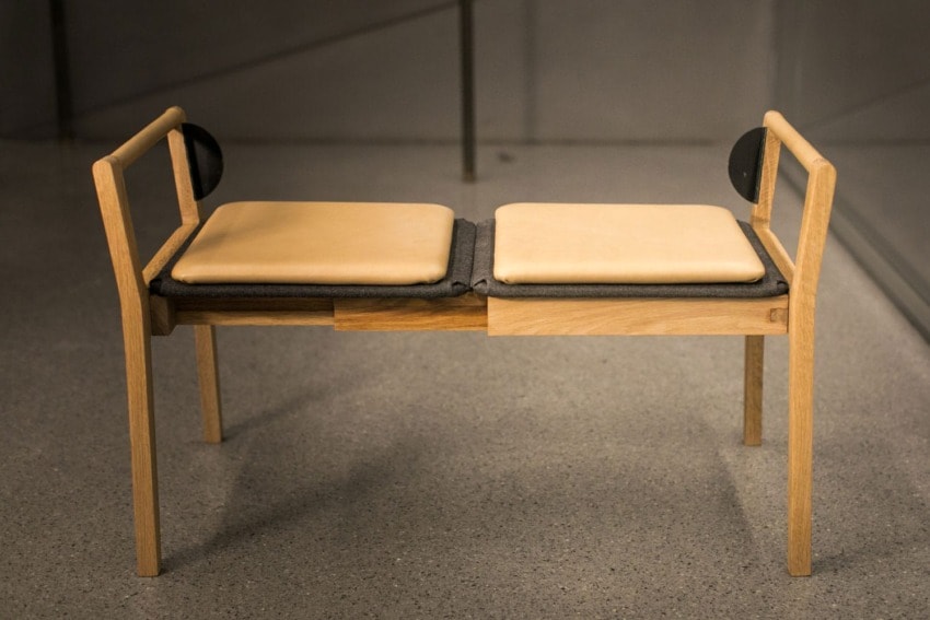 Modern wooden bench with two cushioned seats and simple design, set on a speckled gray floor.