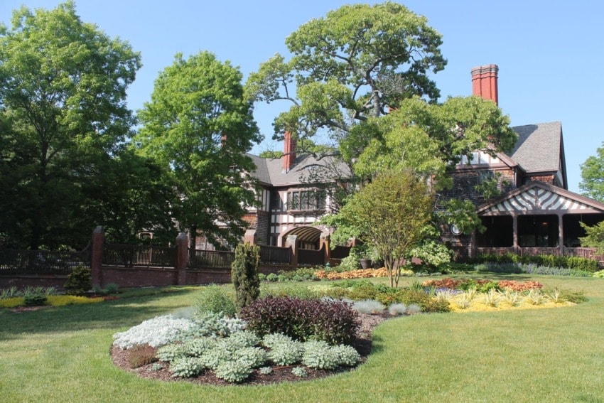 Historic house with lush garden and colorful flowers surrounded by tall trees on a sunny day.