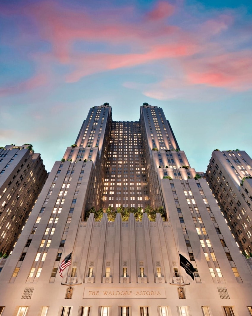 Art Deco-style tall hotel building with illuminated windows and flags, under a sunset sky with colorful clouds.