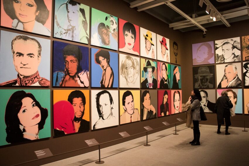 People viewing a gallery wall filled with colorful portraits in a museum setting