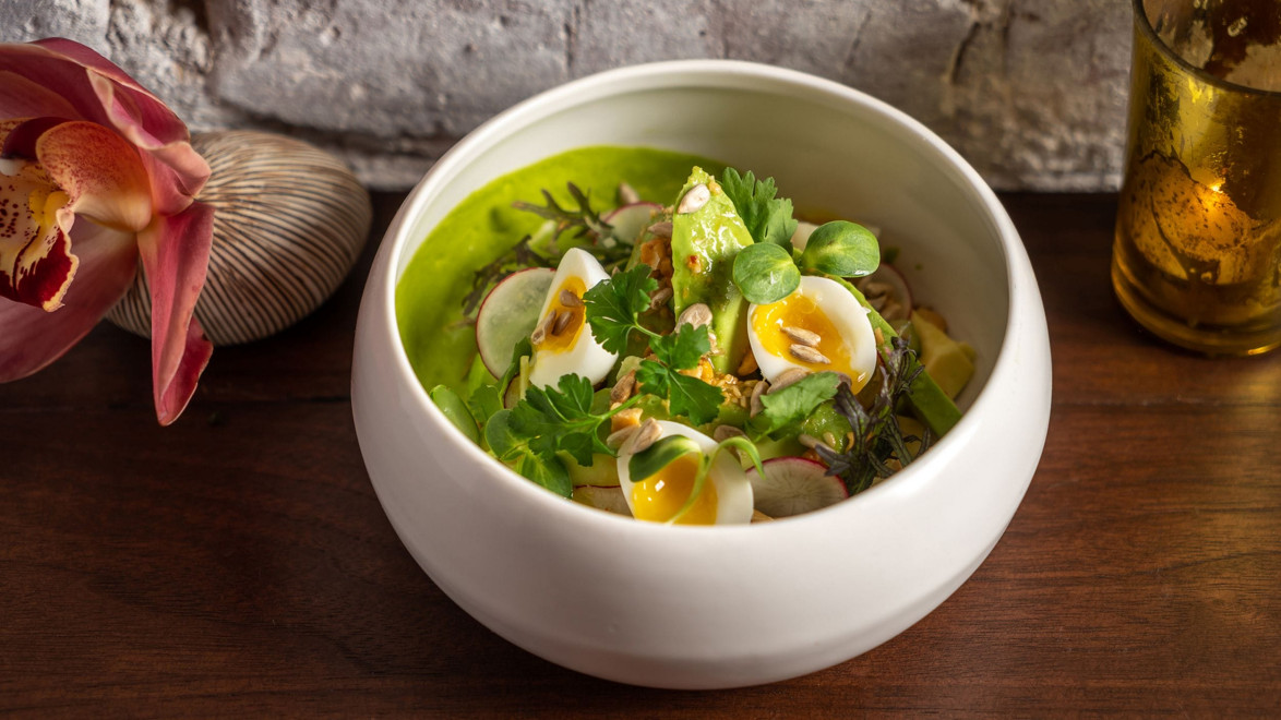 Bowl of green soup garnished with sliced eggs, herbs, and vegetables on a wooden table, next to a flower and candle.