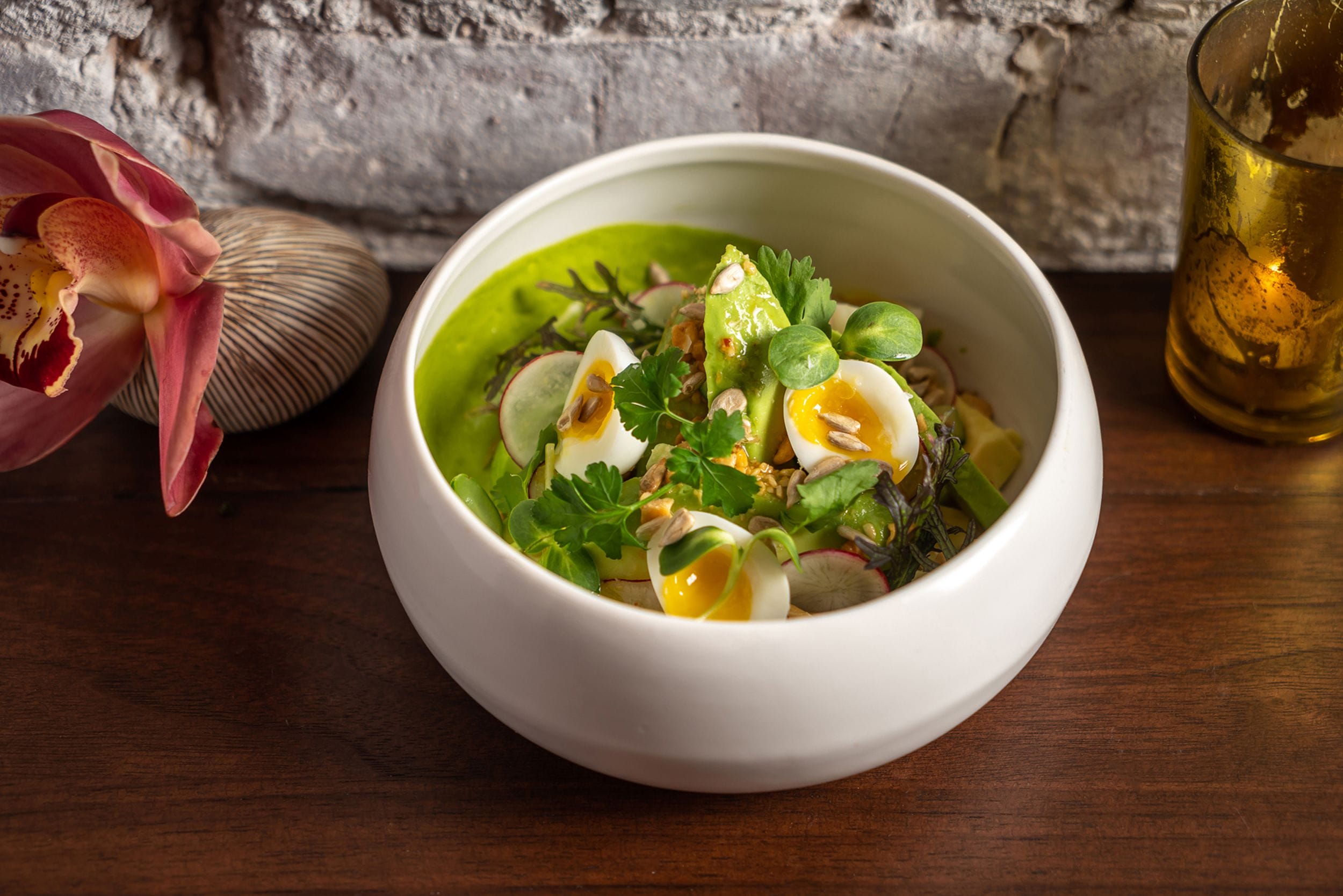 Bowl of green soup garnished with sliced eggs, herbs, and vegetables on a wooden table, next to a flower and candle.