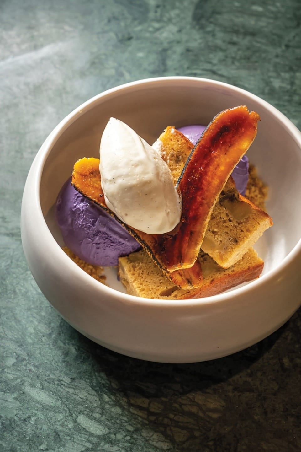 Bowl of ube ice cream with banana slices, cake pieces, and a dollop of cream on a marble surface.