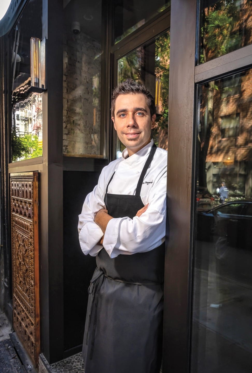 Chef standing outside a restaurant wearing a white chef's coat and black apron, arms crossed, smiling.