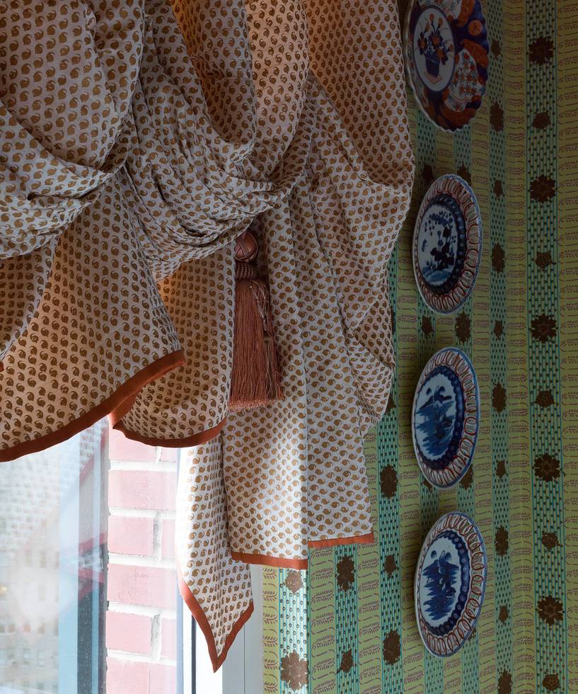 Patterned curtains with tassel draped by a window beside decorative wall plates.