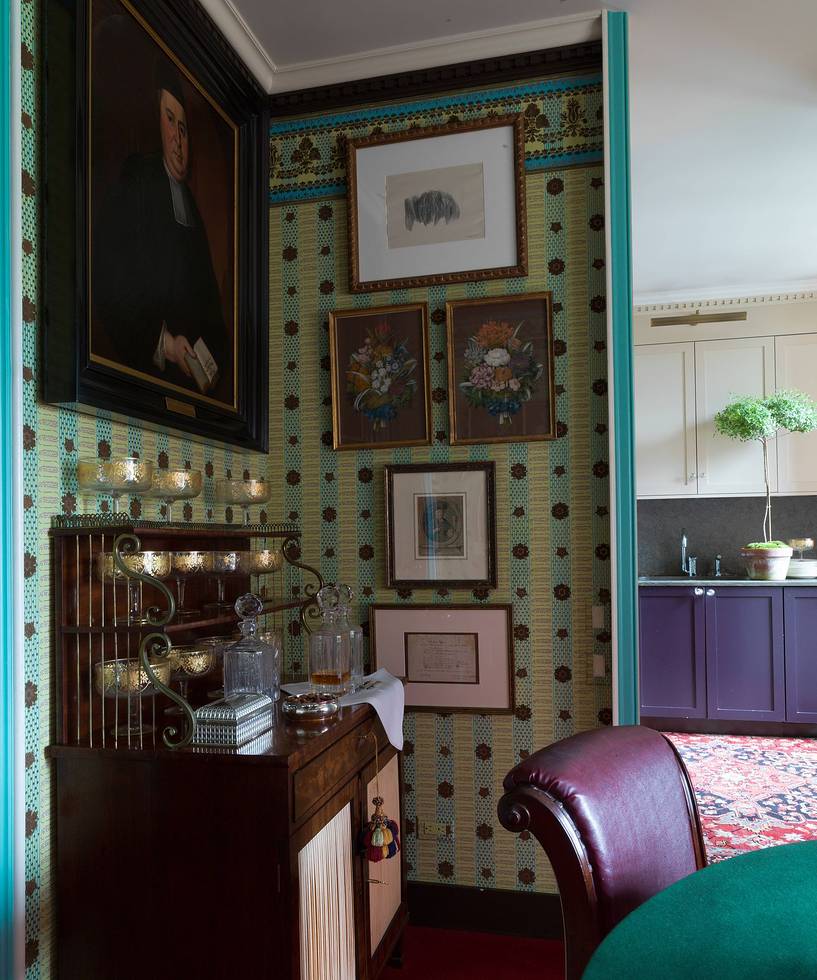 Vintage-style room with patterned wallpaper, framed art, antique furniture, and a glimpse of a kitchen in the background.