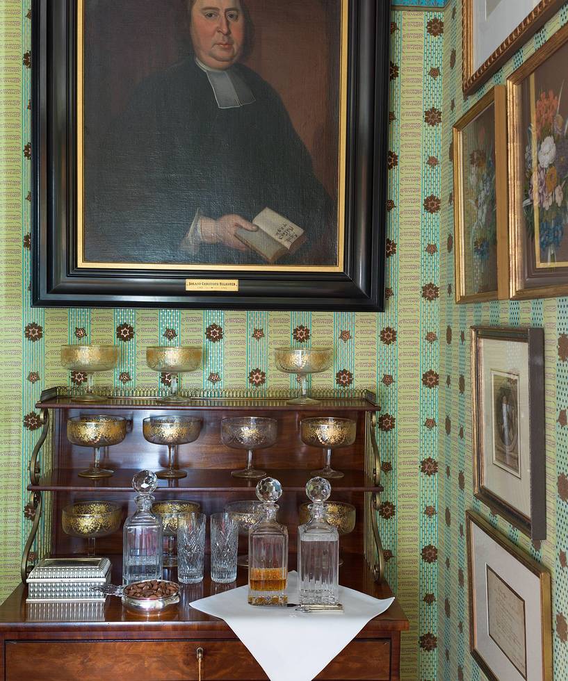 Vintage room with a wooden cabinet, crystal decanters, and silver goblets beneath a historical portrait on patterned wallpapered walls.