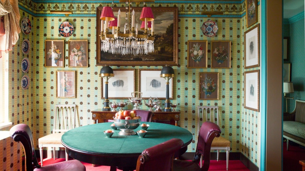 Elegant dining room with patterned wallpaper, green tablecloth, red chairs, chandelier, and framed art on walls.