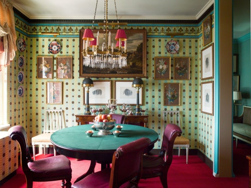 Elegant dining room with patterned wallpaper, green tablecloth, red chairs, chandelier, and framed art on walls.