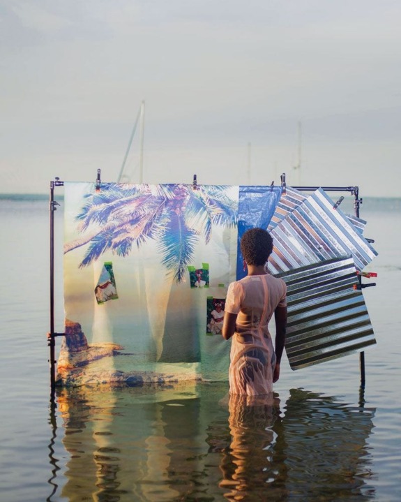 Person standing in water, looking at a display of tropical-themed fabric and metal sheets, with a calm sea background.