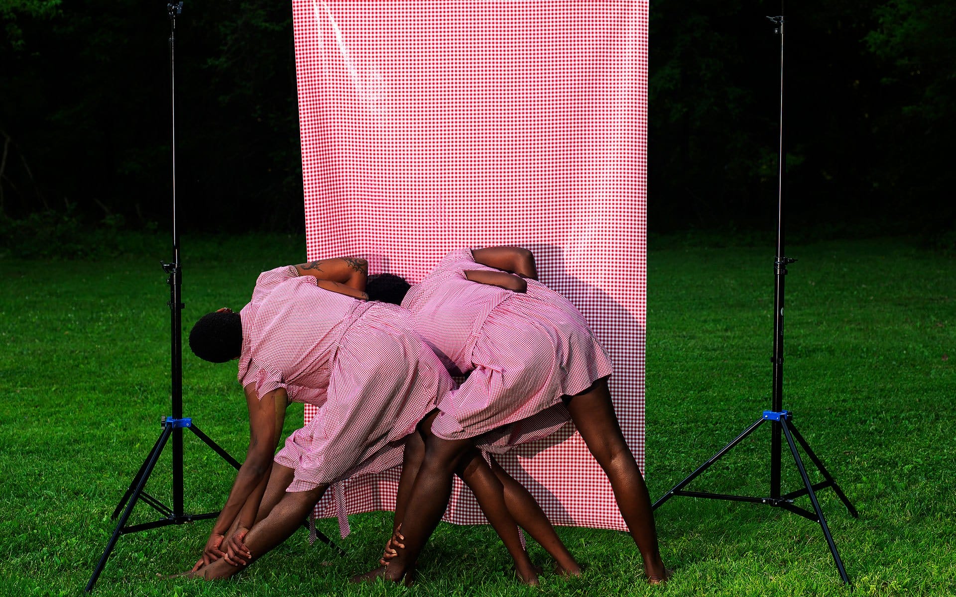 Three people in matching pink checkered dresses bending over in front of a matching backdrop on a grassy field.