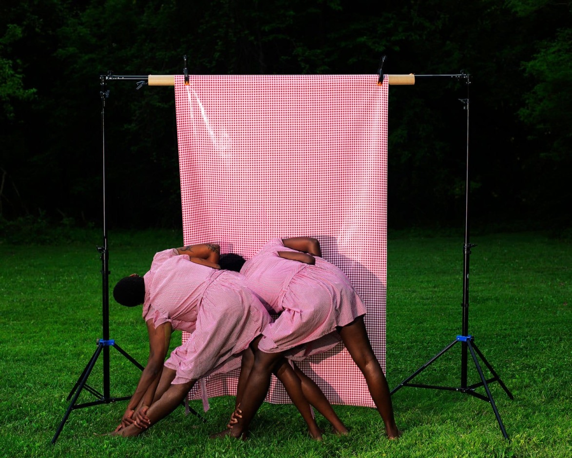 Two people in matching pink dresses pose creatively in front of a pink checkered backdrop on a grassy field.