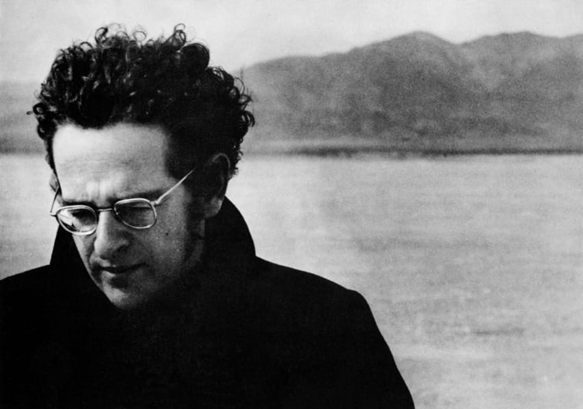 Person with curly hair and glasses outdoors, looking down with mountains in the background. Black and white photo.