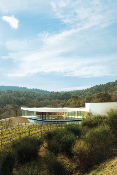 Modern glass building with flat roof surrounded by lush greenery and hills under a blue, partly cloudy sky.