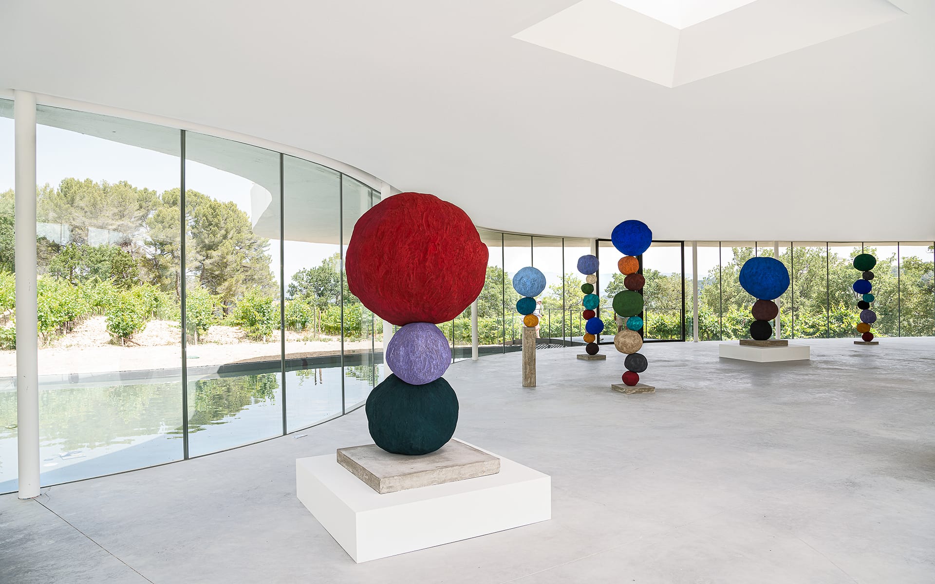 Colorful spherical sculptures displayed in a modern, glass-walled exhibition space with a view of greenery outside.