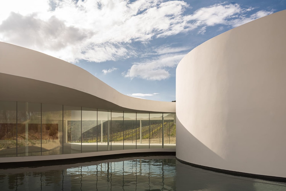 Modern architectural structure with curved white walls, glass windows, and reflections in water under a partly cloudy sky.