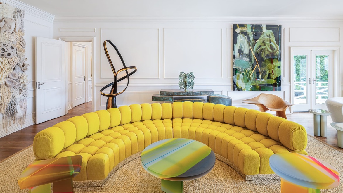 Colorful modern living room with a large yellow curved sofa, abstract art, and vibrant round coffee tables on a woven rug.