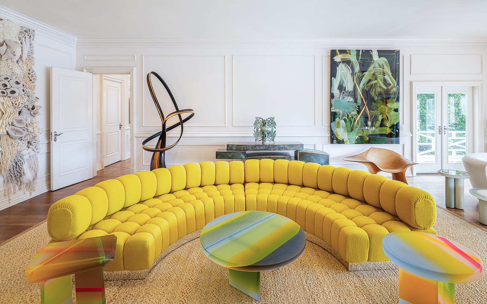 Colorful modern living room with a large yellow curved sofa, abstract art, and vibrant round coffee tables on a woven rug.