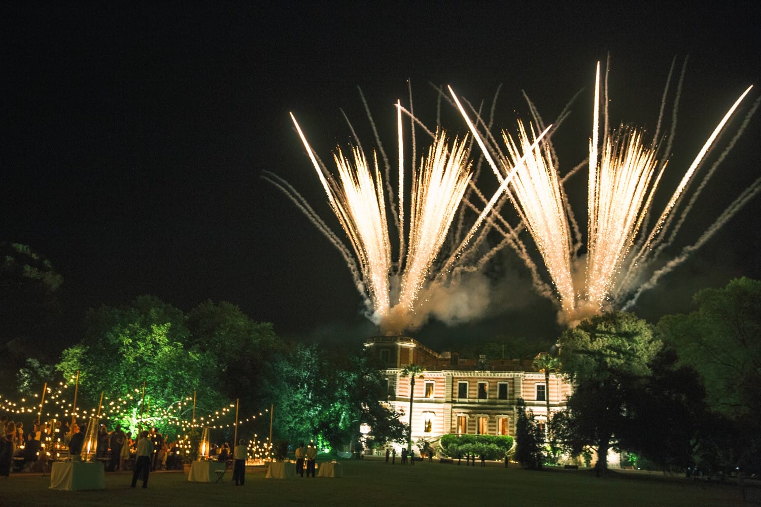 Mansion illuminated at night with fireworks in the sky and festive decorations on the lawn in the foreground.