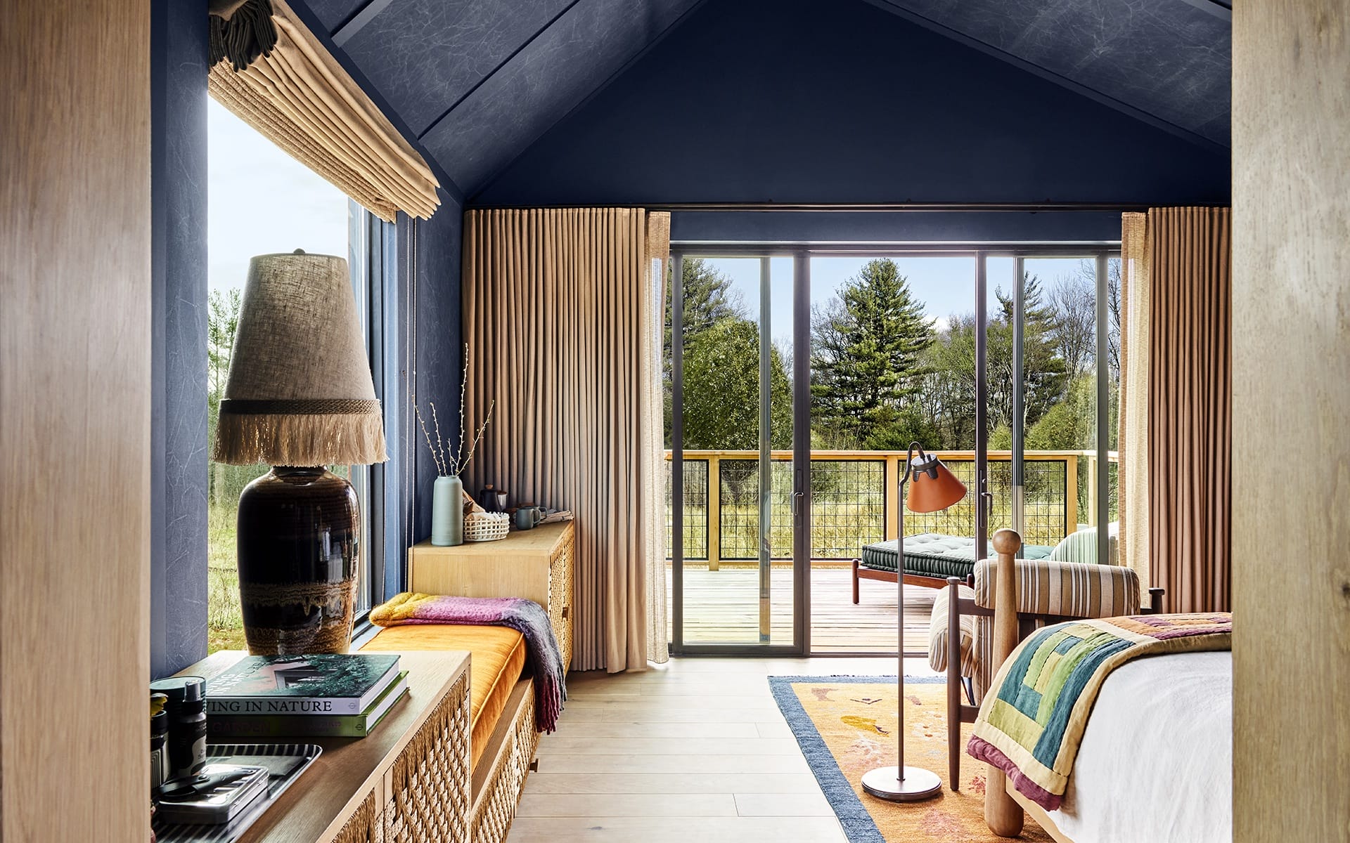 Cozy bedroom with large windows, beige curtains, a colorful rug, and a deck view of greenery outside.