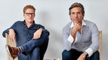 Two men sitting in chairs against a neutral background, one in a blue shirt and the other in a striped shirt, posing thoughtfully.