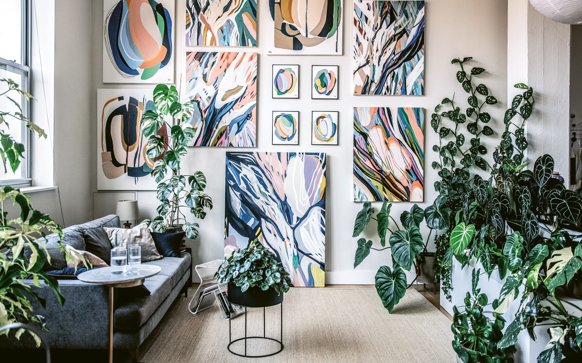 Modern living room with abstract art on walls and lush green plants surrounding a gray sofa and small table.