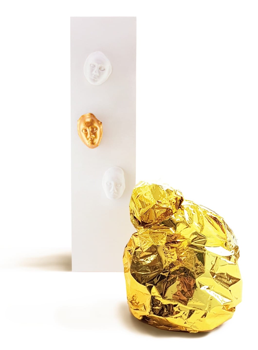 Abstract art installation with three sculpted faces mounted on a white panel and a gold foil-wrapped object in the foreground.