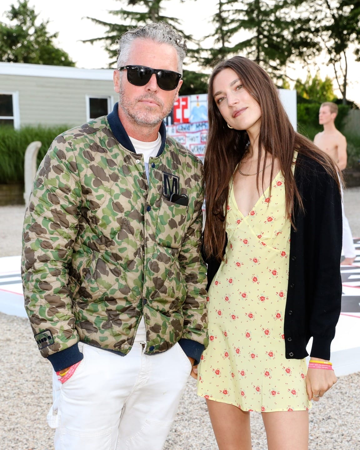 Man in camo jacket and sunglasses with woman in yellow dress and black cardigan standing outdoors on a gravel surface.