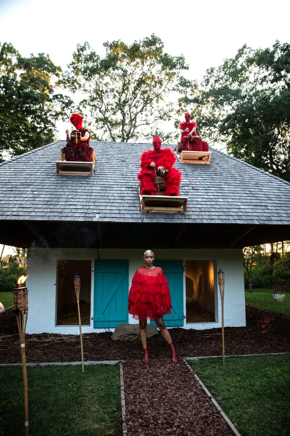 Person in red outfit standing in front of white house with blue doors, three figures in red seated on the roof.
