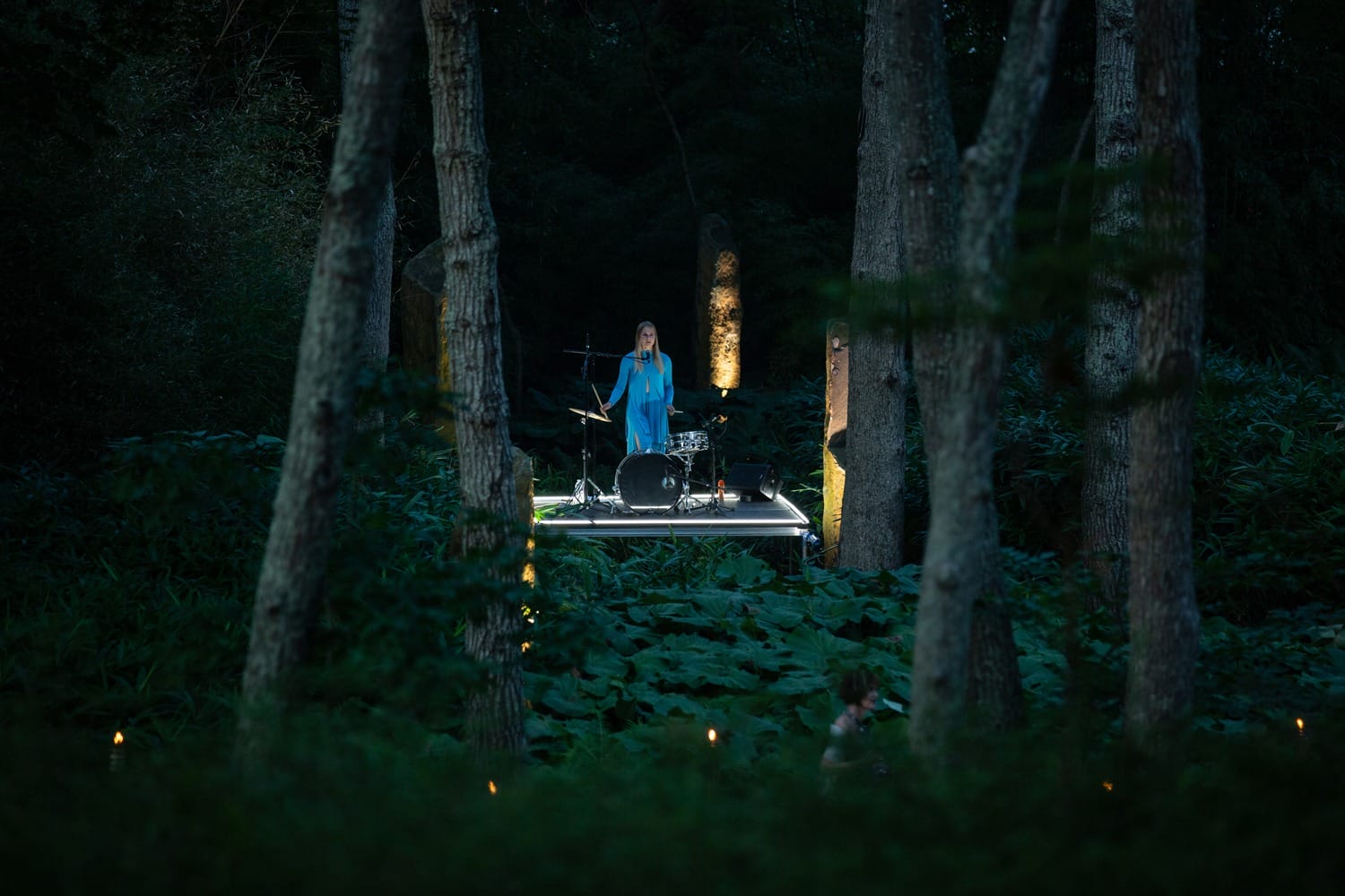 Woman in blue dress sings on an outdoor stage with a drum set, surrounded by tall trees in a dimly lit forest setting.