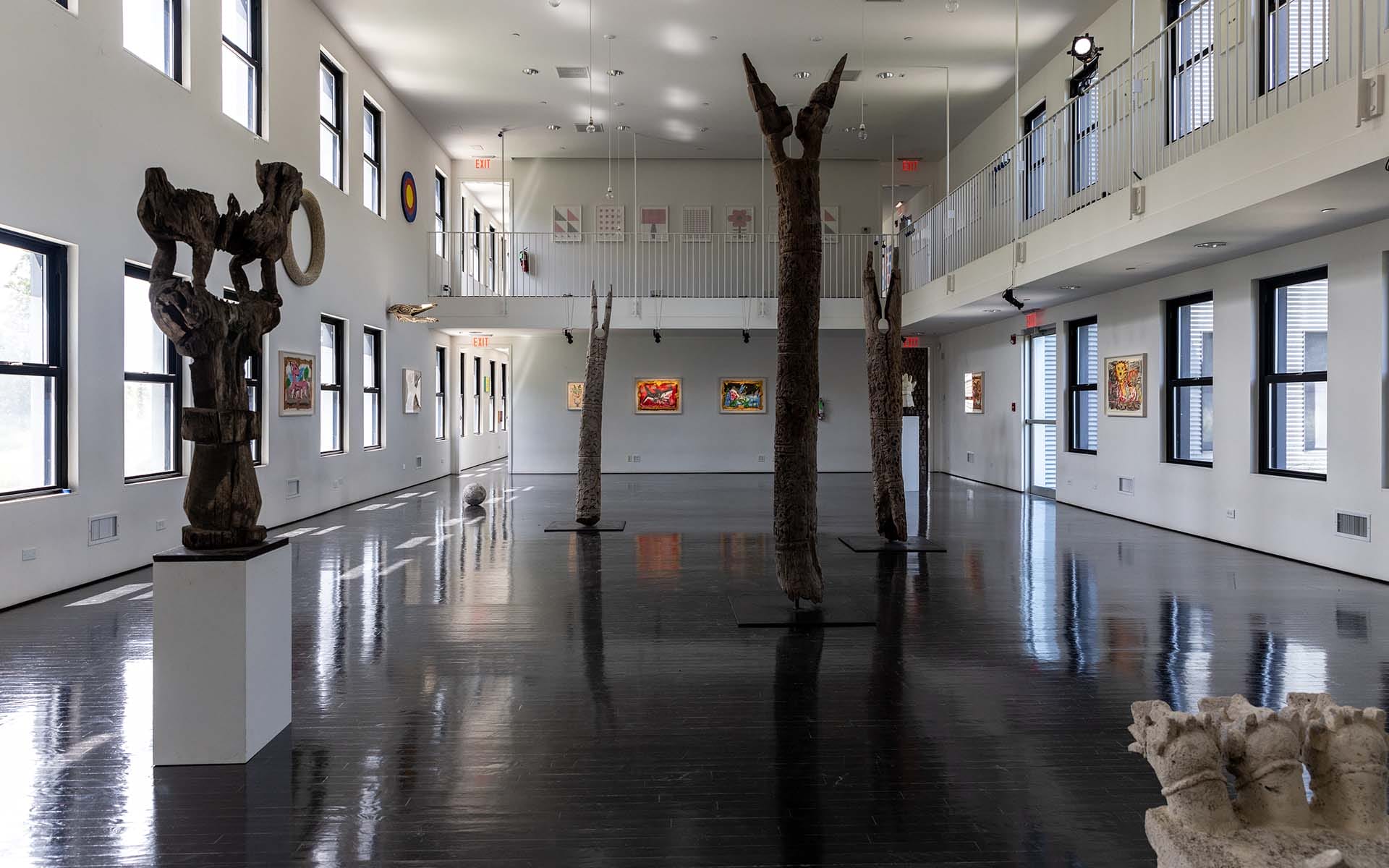 Spacious art gallery with sculptures and colorful paintings on white walls, featuring large windows and dark wooden floors.
