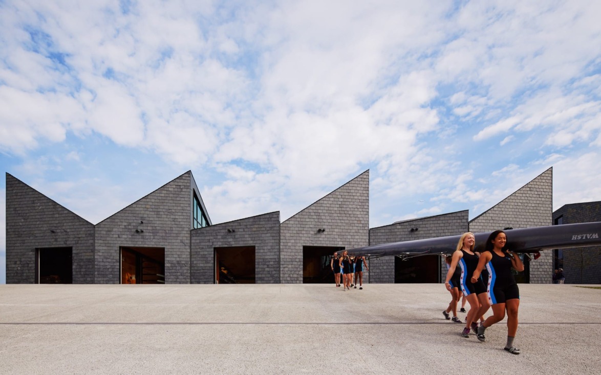 Team of women carrying a racing shell in front of modern boathouse with triangular roofs under a partly cloudy sky