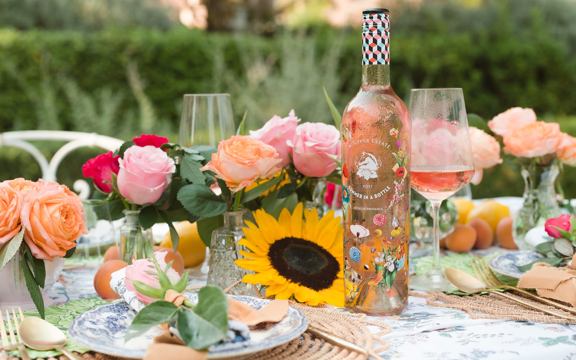 A decorated wine bottle on a floral-themed outdoor table setting with colorful flowers and fresh fruit.