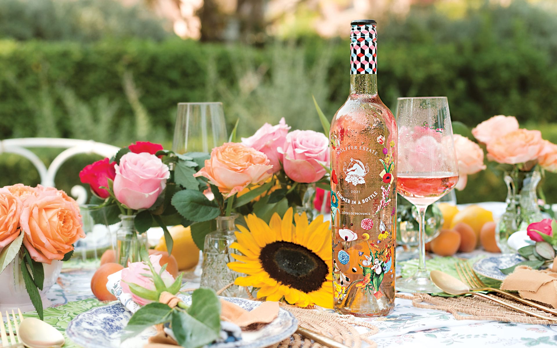 Wine bottle on a floral outdoor table setting with roses and sunflowers in the background.