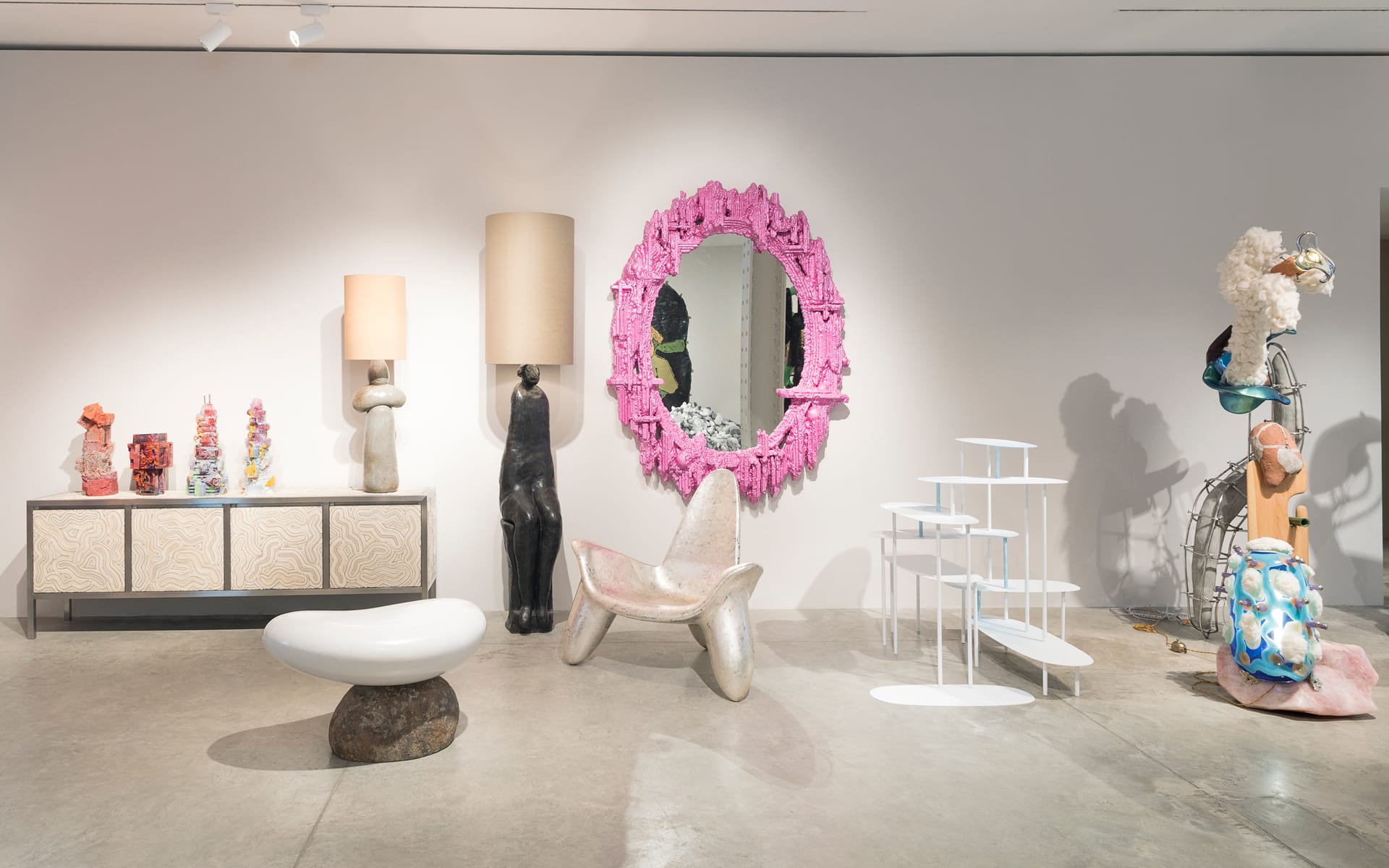 Art gallery interior with eclectic furniture, including a pink framed mirror, abstract sculptures, and various modern decor pieces.