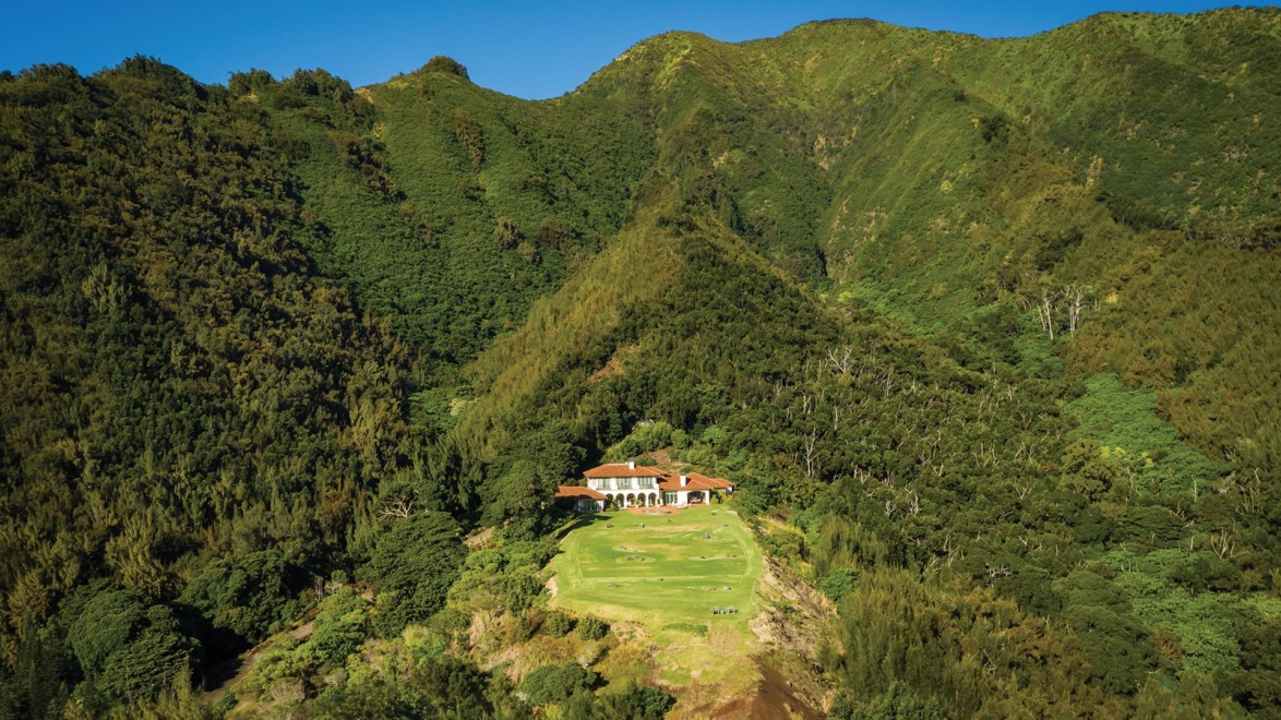 Aerial view of a house nestled in lush green mountains with clear blue sky in the background.