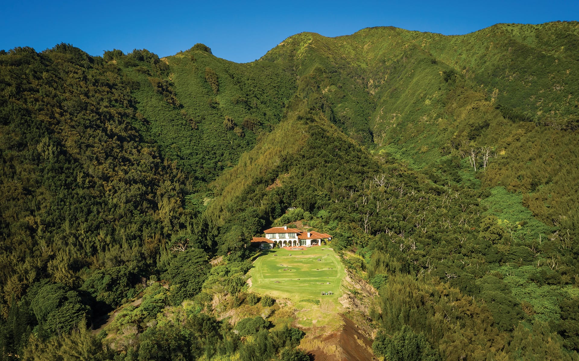 Aerial view of a house nestled in lush green mountains with clear blue sky in the background.