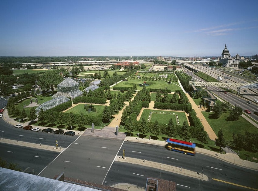 Aerial view of a formal garden with geometric hedges, a glasshouse, surrounding roads, and a cityscape in the background.