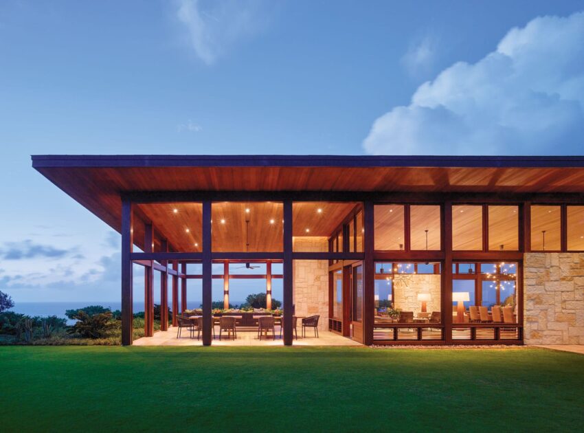 Modern house with large glass windows and wooden roof, overlooking a grassy lawn and cloudy evening sky.