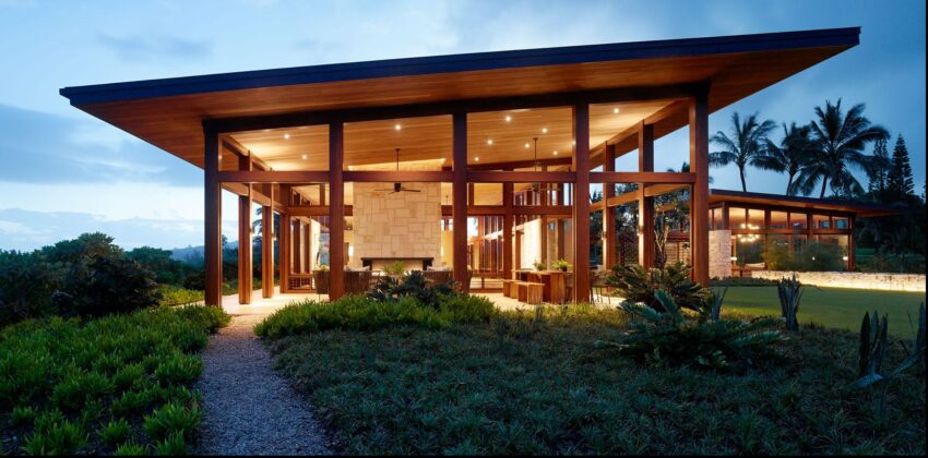 Modern glass and wood house with large windows, lit warmly at dusk, surrounded by lush greenery and palm trees.