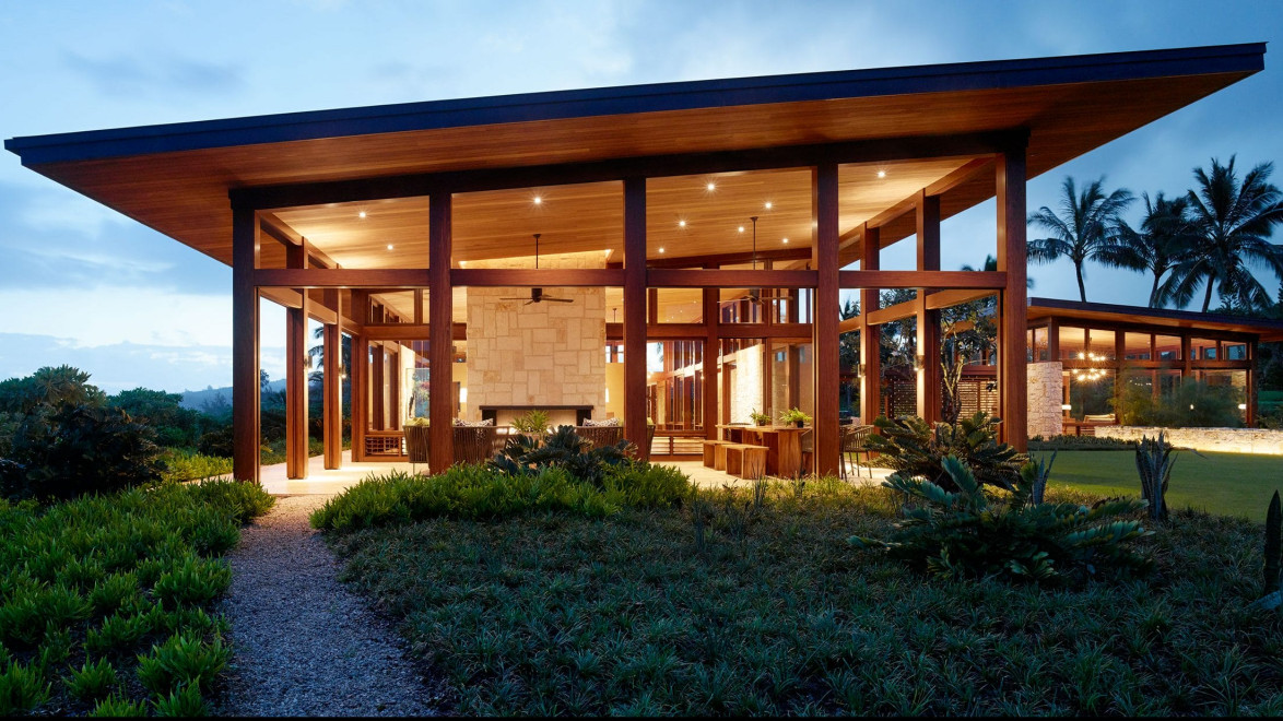 Modern glass and wood house with large windows, lit warmly at dusk, surrounded by lush greenery and palm trees.