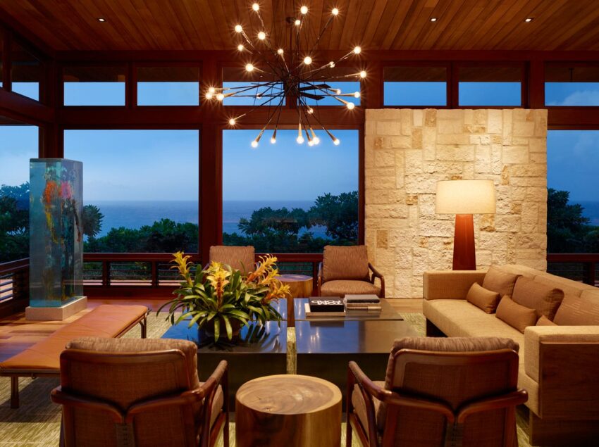Modern living room with large windows, ocean view, cozy seating, chandelier, stone wall, and potted plants.
