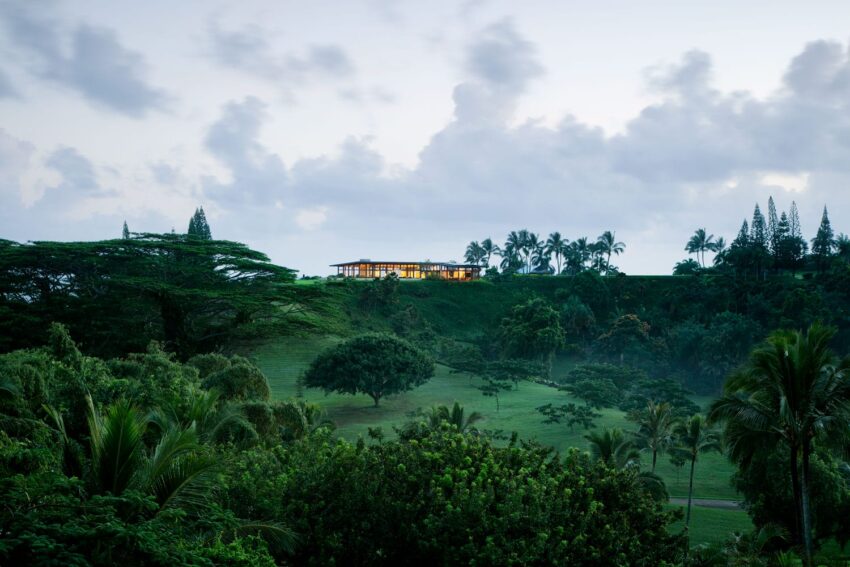 Modern house atop a lush green hill surrounded by tropical trees under a cloudy sky at dusk.
