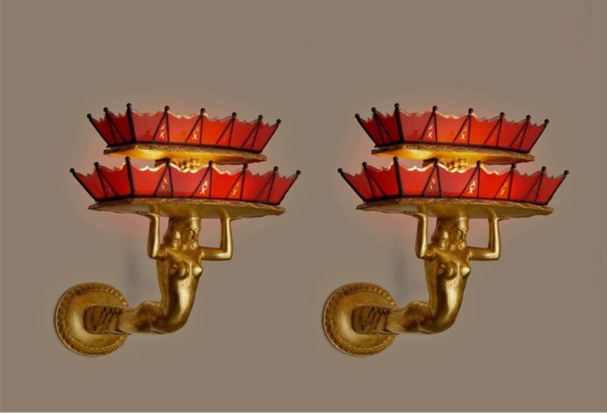 Two ornate wall sconces with golden figures holding red and gold decorative lampshades against a beige background.