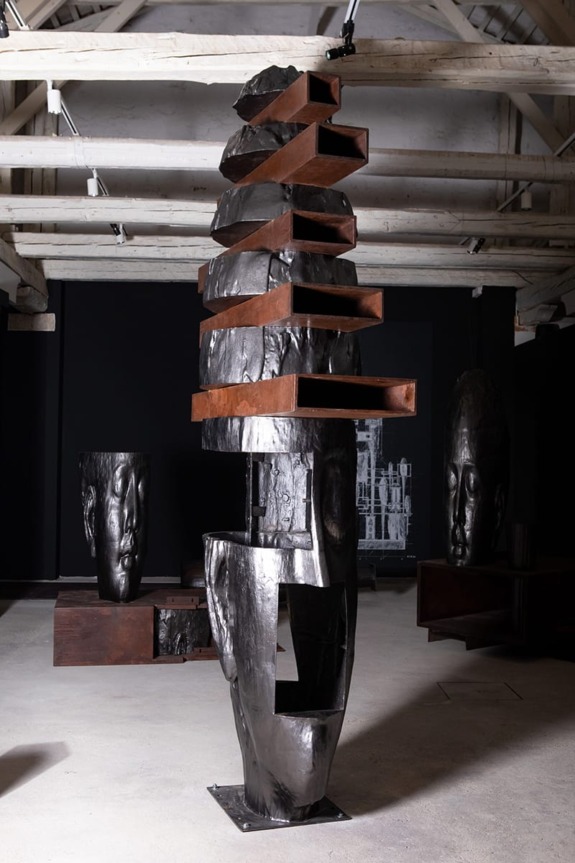 Abstract vertical sculpture with stacked, irregular dark shapes in an art gallery setting with two large face sculptures nearby.