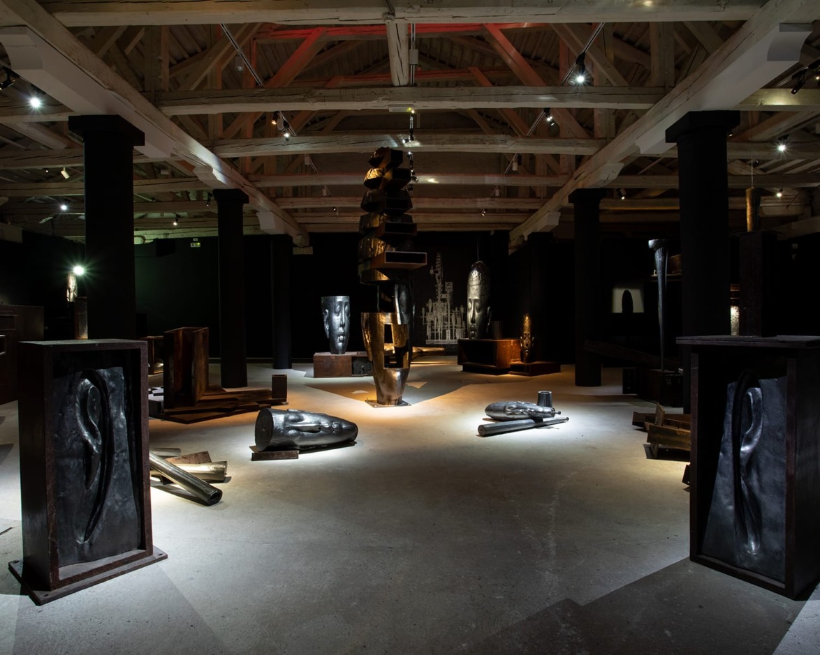 Dimly lit art gallery featuring metal sculptures and head-shaped artworks under a high wooden ceiling.