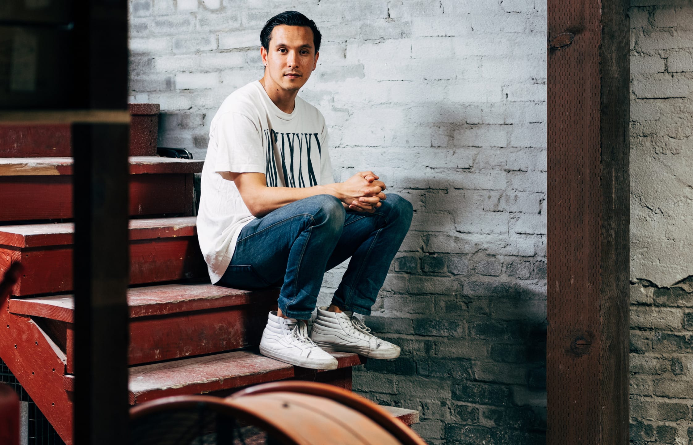 Man in a white t-shirt and jeans sitting on red stairs against a textured brick wall.