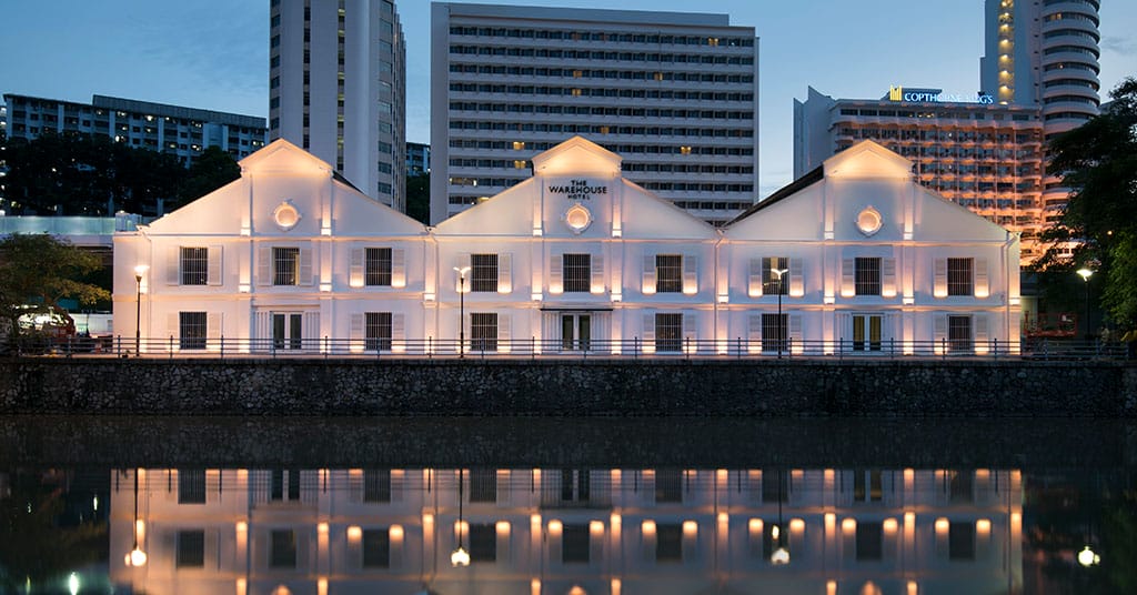 Historic riverside warehouse building lit up at night, reflecting elegantly on calm water with urban skyline in the background.